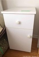 Front view of white laminate bedside table showing drawer and cabinet door with round white knobs. Veneer lifting visible on edge.