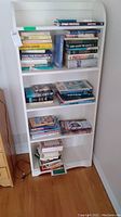White bookshelf with five shelves filled with a variety of books mostly related to sailing, yachting, war, and lifestyle. Books are stacked both vertically and horizontally.