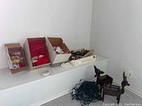 Boxes of assorted Christmas ornaments, string of green lights, wooden reindeer figurines on white ledge against wall.