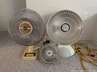 Photo showing three electrical appliances including a beige Home Line oscillating fan, a small gray clip-on fan, and a white Presto Heat Dish heater with metal grill and yellow power cord, all placed on floor against wall.