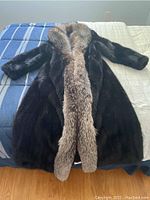 Full view of long black mink fur coat laid flat on bed showing front with raccoon fur collar and trim.