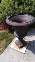 Cast iron planter urn shown from right side, placed on stone tiles with surrounding bushes. Visible wear and rust inside.