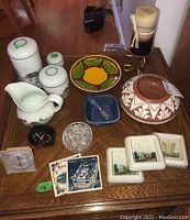 Photo of various pottery bowls, ceramic jug, decorative plates, coasters, crystal glass container, two small clocks, and a tall candle on a metal stand arranged on a wooden table.