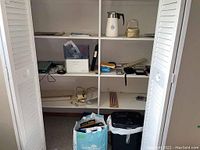 Photo showing white shelving unit with binders, cribbage board, various papers and small items, trash bin, and paper shredder on floor.