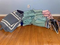 Photo of stacked blue and pink towels, a crocheted blanket, blue soft blankets, and assorted plastic hangers on the floor.