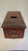 Front view of a rectangular wooden antique cash drawer with a slot on top and brass handle on the front drawer, with visible wear.