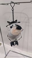 Front angle showing smiling cow ornament hanging by chain and hook on metal stand, ring hanger visible.