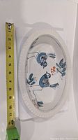 Oval serving dish lying flat with measuring tape showing length approx 11.5 inches. Bluebirds and foliage hand painted in blue and orange colors on dish surface.