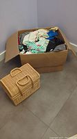 Wide shot showing a large cardboard box filled with assorted quilting fabrics and a vintage wicker sewing basket positioned in front on the floor.