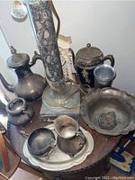 Overview of all brass items on wooden table including coffee pot, teapot, creamer, sugar bowl, decorative bowl, and clock under a lamp.