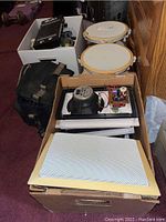 Four Yamaha ceiling speakers stacked in cardboard box with metal grilles visible on top and crossover components visible inside speakers. Black soft vape camera bag behind box.