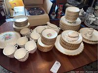 Photo showing stacked plates, bowls, tea cups, saucers, and serving dishes with gold rim and floral design on a wooden table.