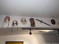 Image showing five ceramic masks and one wall-mounted saber arranged horizontally on a wall.