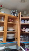 Upper kitchen cupboard filled with stacked plates, bowls, and mugs including Elvis themed item on top