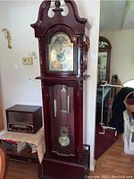 Full view of tall grandfather clock standing against a wall with wooden case and glass front door.