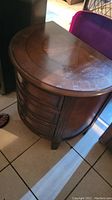 Front and top view of a wooden semi-circular side table on tile flooring showing three drawers with wooden knobs and some dust on surface.