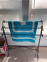 Front view of black metal blanket rack with turquoise and white crocheted blanket hanging on it, positioned in front of window blinds.