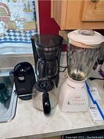Photo showing black Black and Decker coffee maker with glass carafe, black electric can opener, and white Sunbeam blender with glass pitcher on a kitchen counter.