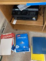 Brother MFC-J435W printer on wooden shelving with multiple packs of printer paper and notebooks below.