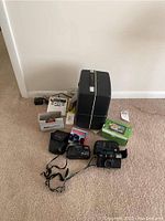 Wide shot shows entire lot, including de Jur Amsco projector, multiple cameras, Garmin device, film rolls, and accessories on carpeted floor next to wall