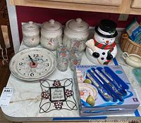 Photo showing three white ceramic canisters with lids, Elgin ceramic wall clock, snowman cookie jar, Marmalade flatware set in box, three drinking glasses, and decorative metal sign.