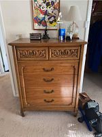 Front view of wooden five drawer chest of drawers with decorative carved details on upper drawers and brass handles.