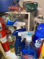 Various household cleaning spray bottles and liquid cleaners on shelf including Rug Doctor upholstery cleaner, and Old English wood cleaner bottle.