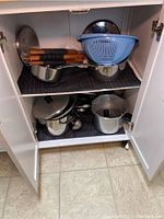 View of kitchen cabinet shelf showing cookware including pots and pans with lids, cooking utensils, and blue plastic strainer