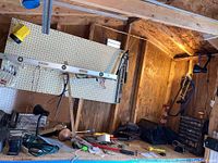 Wide-angle view of workbench and pegboard in shed with tools and equipment including levels, electrical cords, and hand tools scattered on bench.