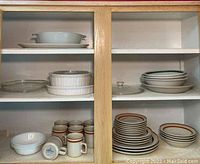 Cabinet shelves displaying white ceramic casserole dishes, glass pie plate, several mugs, bowls, and plates with brown and green trim.