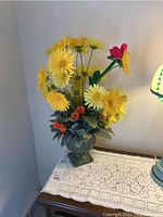 Full view of yellow and red faux flower arrangement in decorative green vase on a lace doily-covered table with part of a lamp visible.
