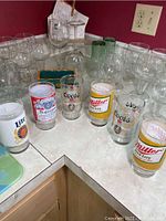 Assortment of beer glasses with Miller Lite, Budweiser, Coors, and Miller logos on a kitchen counter