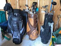 Three golf bags with clubs inside, some clubs visible with head covers, bags against wood wall and concrete floor.