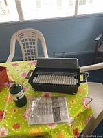 Portable hibachi grill on floral tablecloth next to green Bernzomatic camping gas canister and manual.