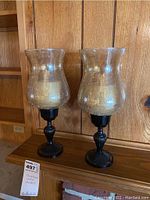 Pair of hurricane candle holders displayed on wooden surface against wood panel wall. Each has black decorative base with glass chimney that has bubbled/frosted texture.