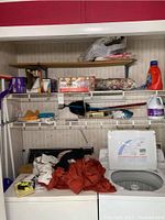 Photo shows garage or laundry area shelves with cleaning supplies and laundry detergent bottles alongside an open top-loading washing machine filled with cloths and items.