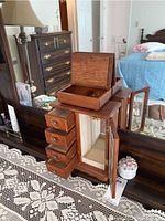 Two wooden jewelry boxes on a table; one box with four drawers and a glass front door is open showing interior necklace hooks and a mirror. The other is a smaller wooden box with the lid open revealing two compartments.