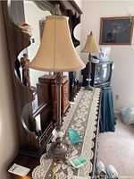 Pair of tall table lamps with beige bell-shaped shades on dark wood dresser with lace runner.