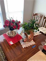 Photo showing faux red flower arrangement in ceramic pot and pitcher bowl set, live green plant in green ceramic pot, and hanging bird-shaped wind catcher on a wooden table near window.