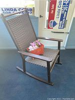 View of woven resin and wood rocking chair with metal planter and faux pink flowers on the seat.