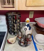 View of metal rotating spice rack with labeled spice bottles, ceramic goose dish with lid, stainless steel condiment caddy with multiple bottles, and ceramic spoon rest on kitchen countertop.