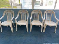 Four white plastic stacking arm chairs arranged side by side on a porch, showing overall view and condition.