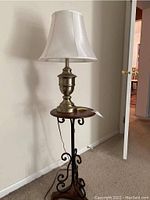 Floor lamp with white fabric shade and brushed silver finish metal body atop a small wooden table with black metal scrollwork base on carpeted floor near white wall and open door.