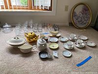 Full lot of teacups, teapots, gravy boats, mugs, and glassware arranged on carpeted floor beneath window with blinds.