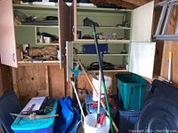 Wide view of shelving with assorted tools, boxes, plastic totes, buckets, and broom leaning against shelf in shed.
