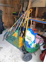 Photo shows assorted yard tools leaning against wall inside shed, including rakes, shovels, garden cart, and additional tools near a workbench.