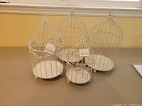 Photo showing 4 decorative white metal bird cages with leaf details and hanging loops, arranged on a table by a window.