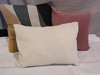 Photo of three accent throw cushions: multicolor gold-grey-white-black striped square cushion, mushroom pink square cushion, and winter white rectangular cushion on a neutral surface.