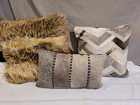Three designer throw cushions arranged together showing textures and patterns: faux fur, faux hide with chevron pattern, and rectangular faux hide with stitched strips.
