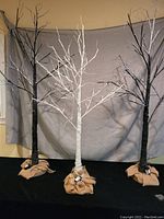 Three rustic light up trees, two black approximately 5 feet tall and one white approximately 4 feet tall all with burlap wrapped bases and multiple branches with integrated lights.